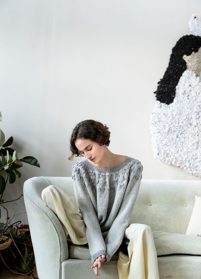 Manaca Sweater: gray knit sweater with patterned neckline and long sleeves, photographed seated on a green sofa with textured wall art and plants.