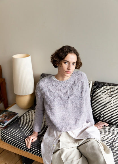 Musgo Sweater: light purple fuzzy knit sweater with long sleeves, photographed seated on a patterned couch beside a lamp.
