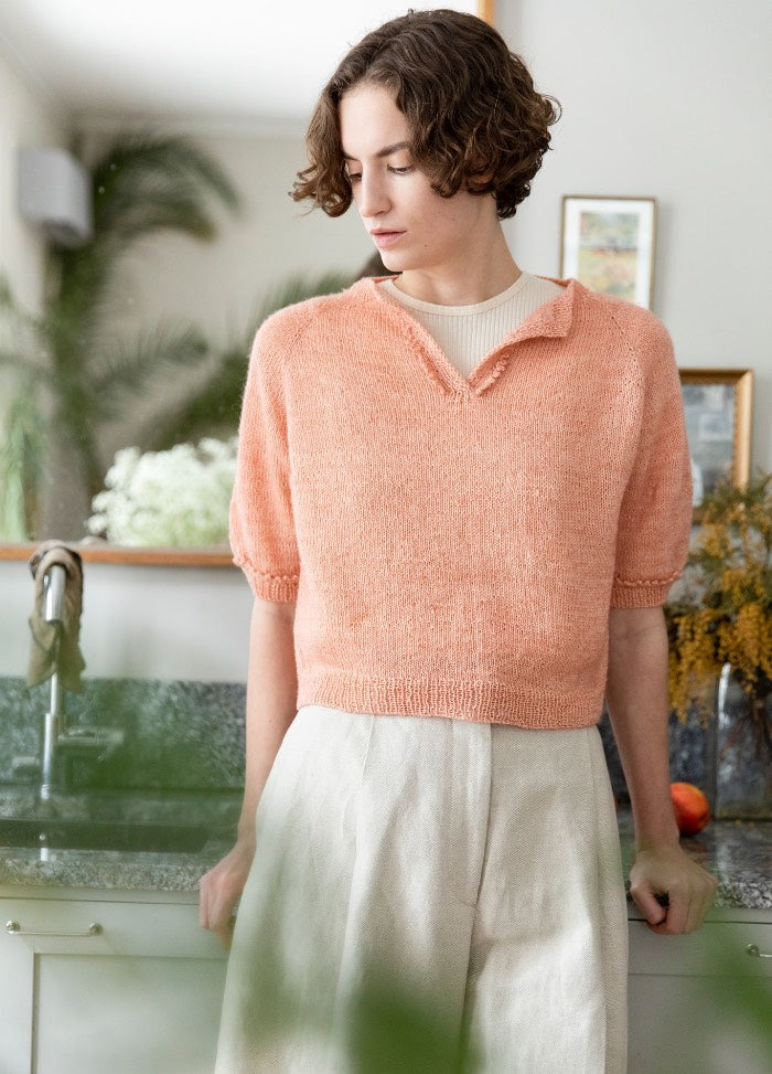 Viana Top: light peach knit top with short sleeves and V-neck, worn with light pants, photographed in a kitchen setting.