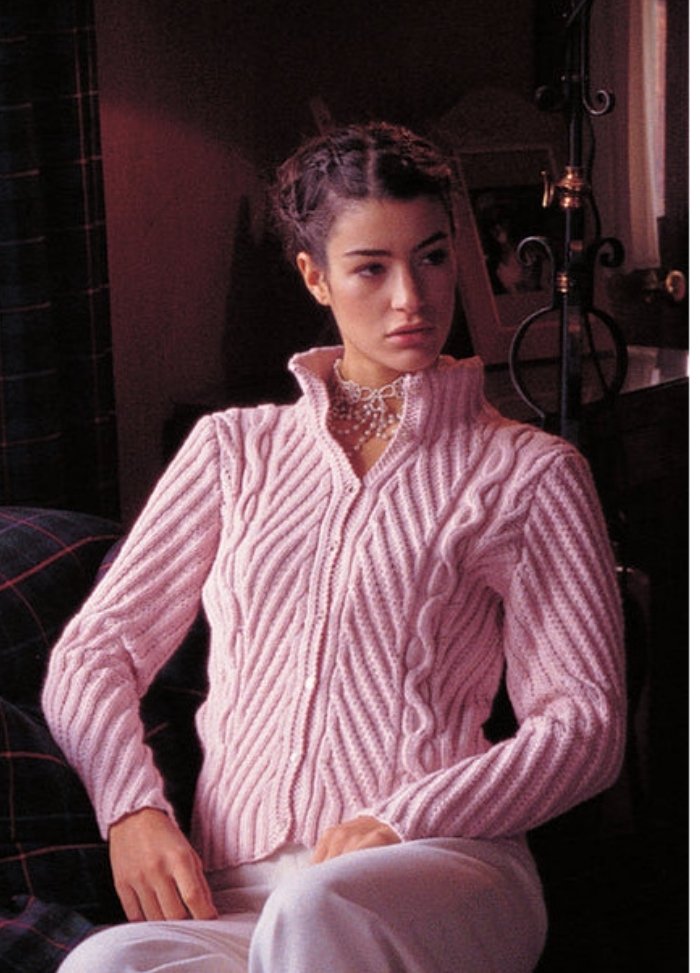 A woman with braided hair wears a pink, cable-knit sweater and white pants, sitting beside a lamp. She appears thoughtful, in a softly lit room.