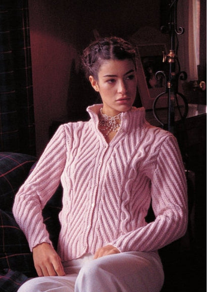 A woman with braided hair wears a pink, cable-knit sweater and white pants, sitting beside a lamp. She appears thoughtful, in a softly lit room.