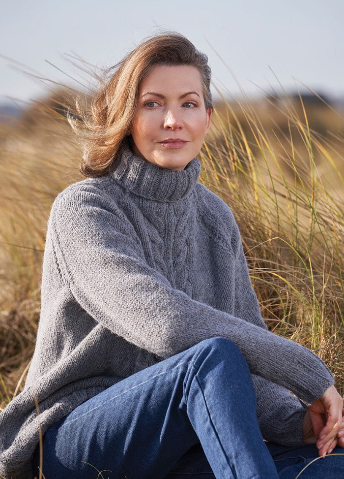 A person in a cozy gray sweater and jeans sits in a serene field of tall grass, with a thoughtful expression. The scene conveys a calm, contemplative mood.