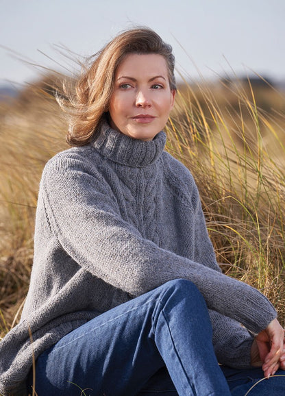 A person in a cozy gray sweater and jeans sits in a serene field of tall grass, with a thoughtful expression. The scene conveys a calm, contemplative mood.