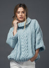 Woman in a cozy, light blue cable-knit sweater with a rolled turtleneck, posing against a gray background, exuding warmth and casual elegance.