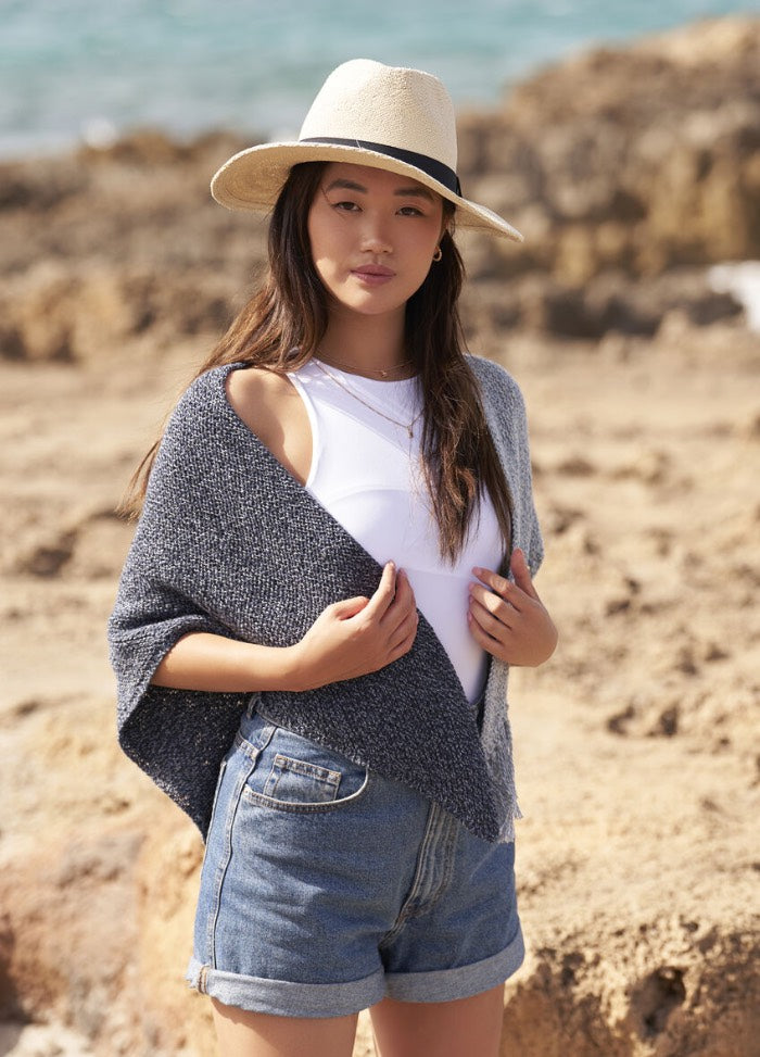 Young woman on a beach, wearing a beige hat, gray shawl, white top, and denim shorts. She stands confidently with a serene ocean background.