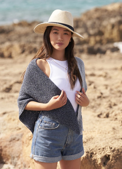 Young woman on a beach, wearing a beige hat, gray shawl, white top, and denim shorts. She stands confidently with a serene ocean background.