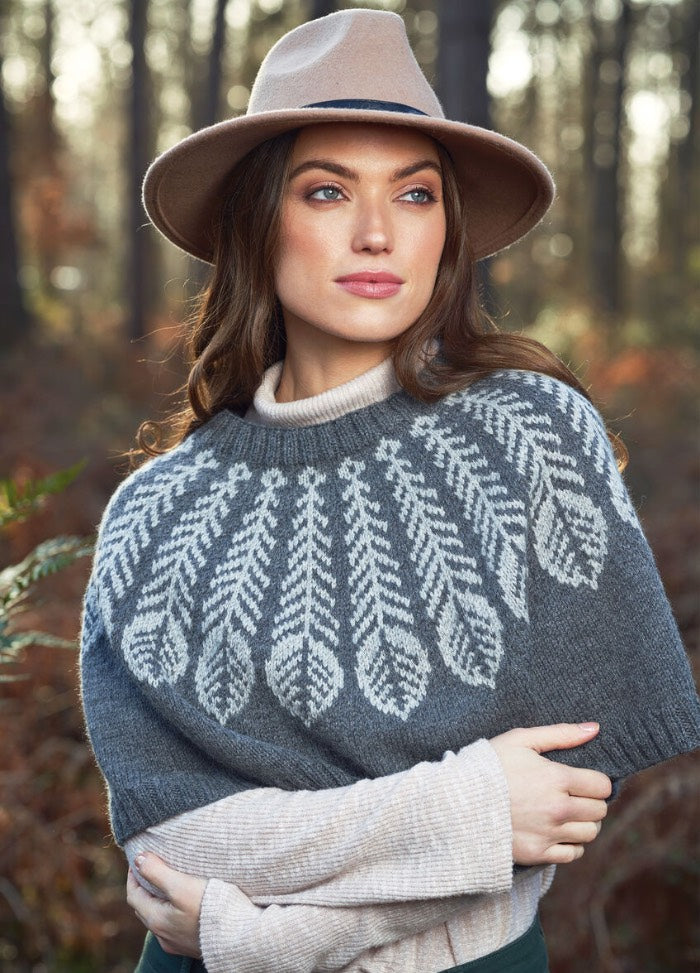 Woman in a gray sweater with intricate white patterns and beige hat stands in a forest, gazing to the side. Autumn leaves create a warm, serene backdrop.