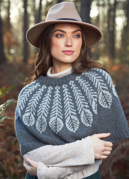 Woman in a gray sweater with intricate white patterns and beige hat stands in a forest, gazing to the side. Autumn leaves create a warm, serene backdrop.