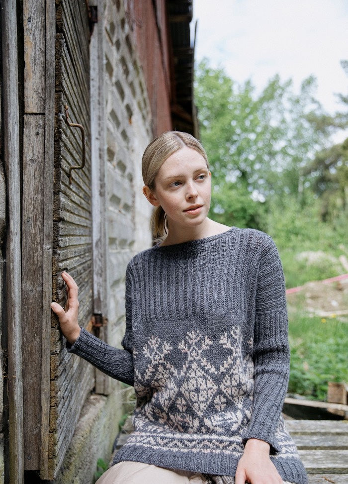 Pohjola Sweater: grey knit featuring white geometric and reindeer motifs along the lower half, worn outdoors near a rustic wooden structure.