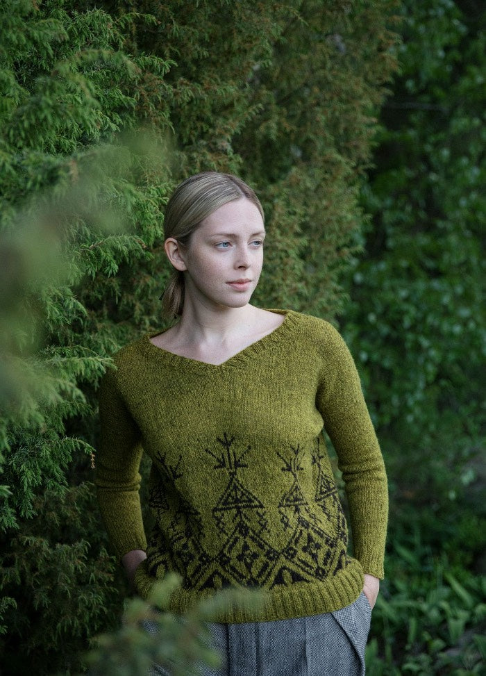 Pohjolan Em?§nt?§ Sweater: green knit with dark decorative motifs on the hem and sleeves, photographed outdoors among lush greenery.