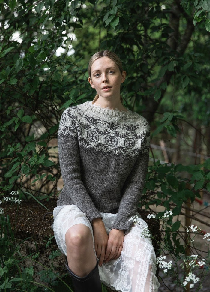 Pohjolan Neito Sweater: grey knit with a white and black yoke pattern, styled with a white lace skirt and black boots in a lush outdoor setting.