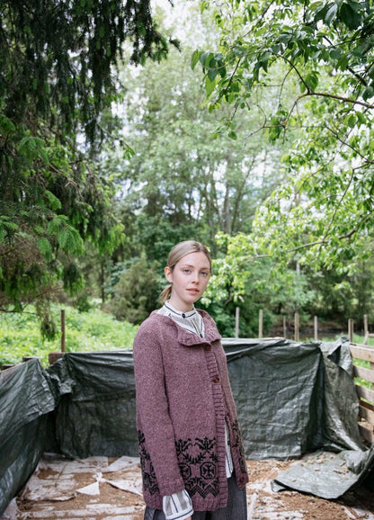 Pohjolan Neito Cardigan: pink knit with black motifs near the hem, worn outdoors among trees and greenery.
