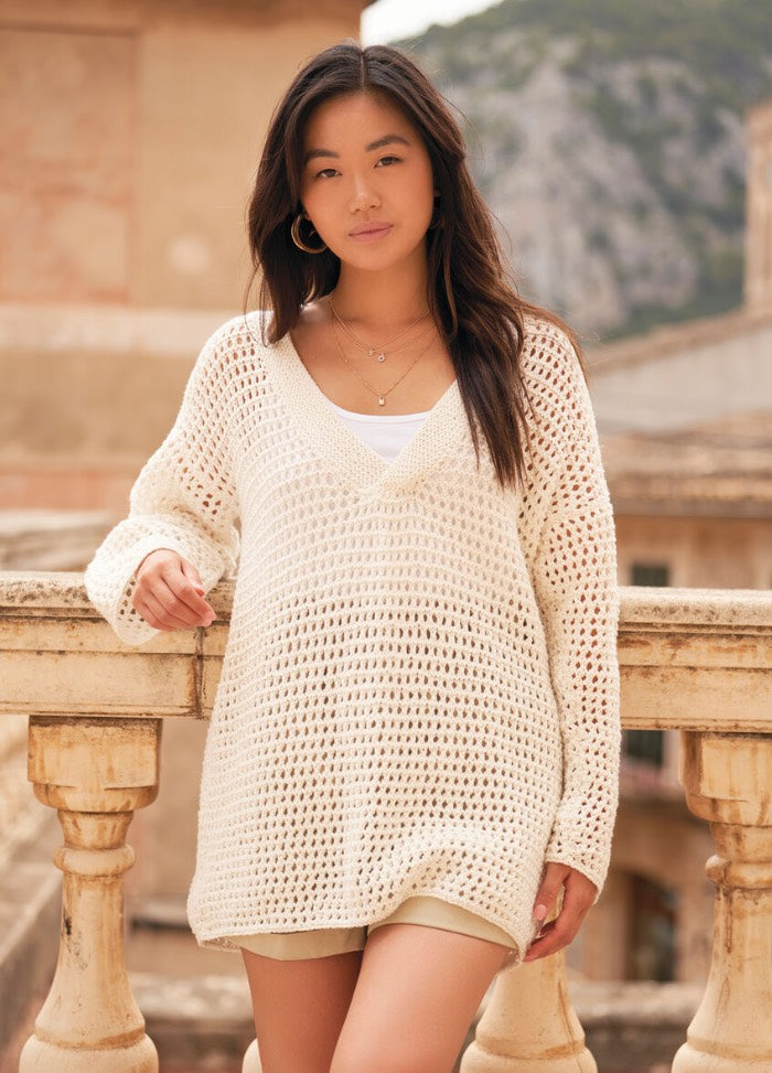 Young woman in a cream crochet sweater leans on a stone balcony, set against a mountainous backdrop. She appears calm and confident.