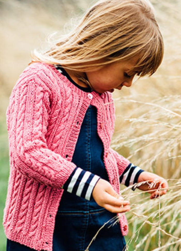 Poppy Cardigan: Young child in a pink hand-knitted cable-pattern cardigan over a dark blue dress with striped sleeves, standing in tall dry grass outdoors.