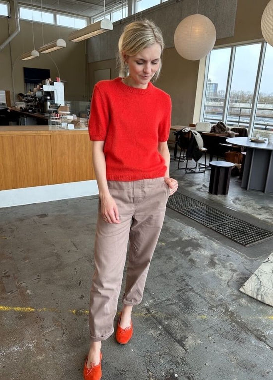 A woman in a red sweater and beige pants stands in a modern café with wood accents and large windows, conveying a relaxed and casual vibe.