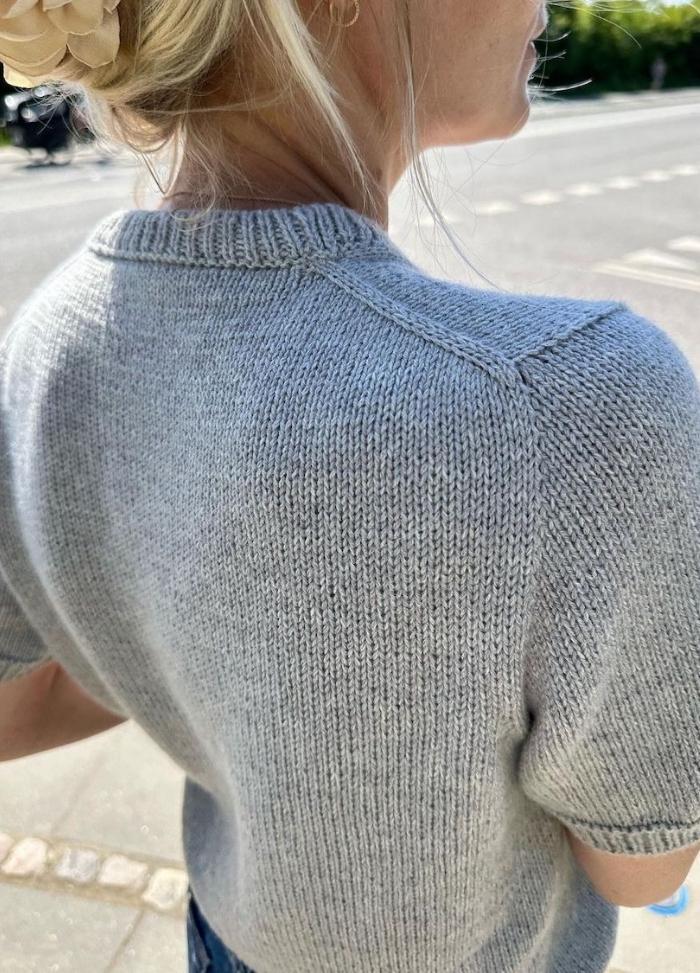 A woman in a gray knitted sweater stands on a sunny sidewalk. Her hair is styled in a bun with a large beige flower accessory. The scene feels casual and relaxed.