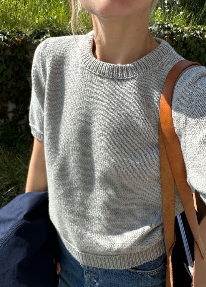 A person in a light gray knit sweater and blue jeans holds a navy jacket and a brown leather bag strap. Sunlight casts soft shadows. Vegetation is visible behind.