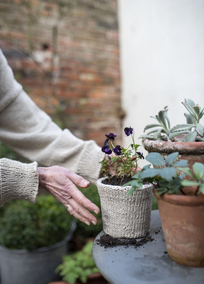 Elderly hands place a plant in a knitted cover on a table, surrounded by pots. The scene conveys a serene, rustic gardening setting.