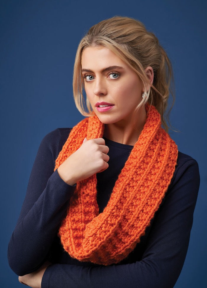 Woman with light hair wearing a bright orange knitted scarf and navy blue sweater poses against a solid blue background, conveying a calm confidence.