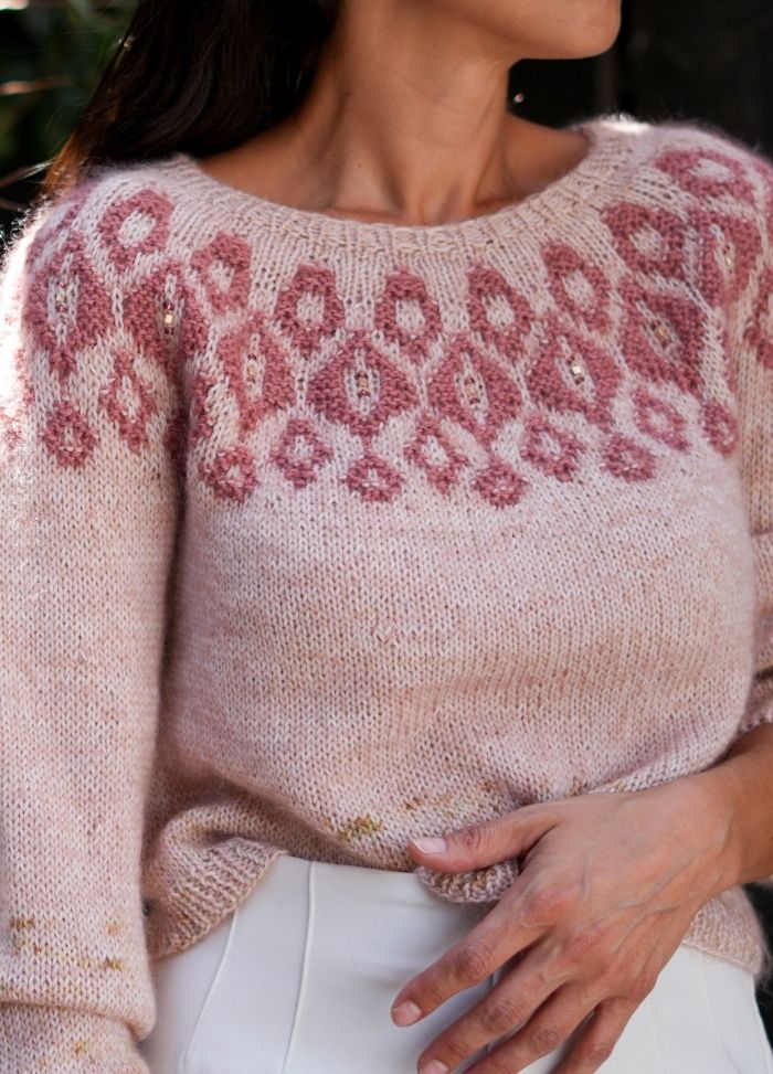 Woman in light pink sweater with intricate red pattern around the neckline. The sweater appears soft and cozy, paired with white pants. The setting is casual and elegant.