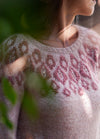 A woman in a soft pink sweater with intricate maroon patterns gazes to the side, partially obscured by green leaves, evoking a serene, contemplative mood.