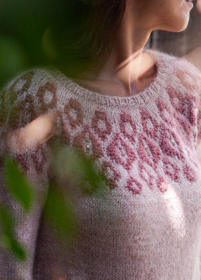 A woman in a soft pink sweater with intricate maroon patterns gazes to the side, partially obscured by green leaves, evoking a serene, contemplative mood.