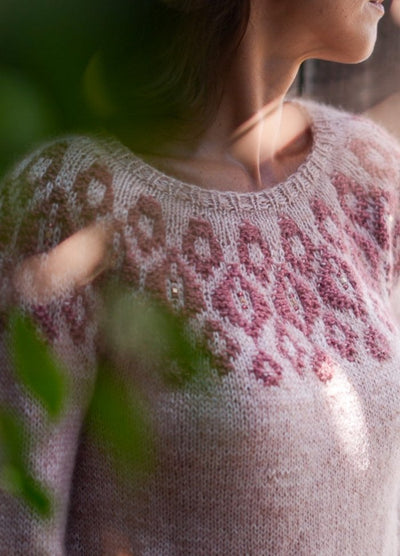 A woman in a soft pink sweater with intricate maroon patterns gazes to the side, partially obscured by green leaves, evoking a serene, contemplative mood.