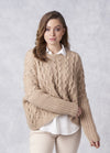 A woman poses against a light gray background, wearing a cozy beige cable-knit sweater over a white shirt. She looks confident and relaxed.