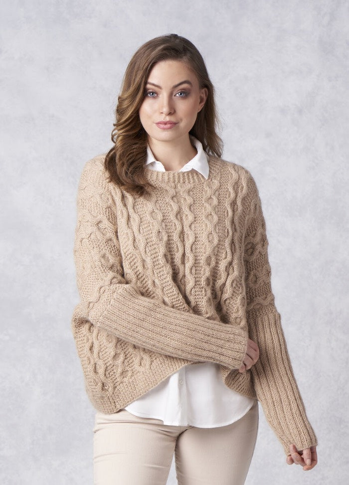 A woman poses against a light gray background, wearing a cozy beige cable-knit sweater over a white shirt. She looks confident and relaxed.