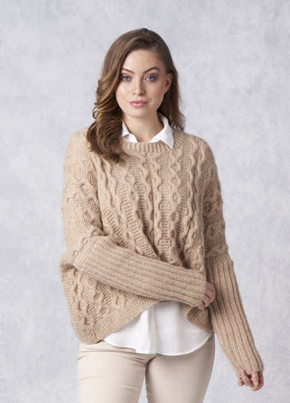 A woman poses against a light gray background, wearing a cozy beige cable-knit sweater over a white shirt. She looks confident and relaxed.