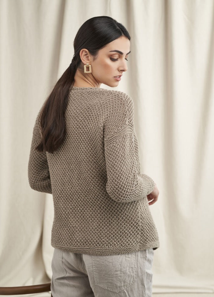A woman with long dark hair, wearing a textured taupe sweater and beige pants, looks over her shoulder against a neutral draped curtain background.