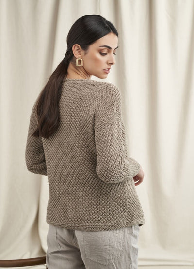 A woman with long dark hair, wearing a textured taupe sweater and beige pants, looks over her shoulder against a neutral draped curtain background.