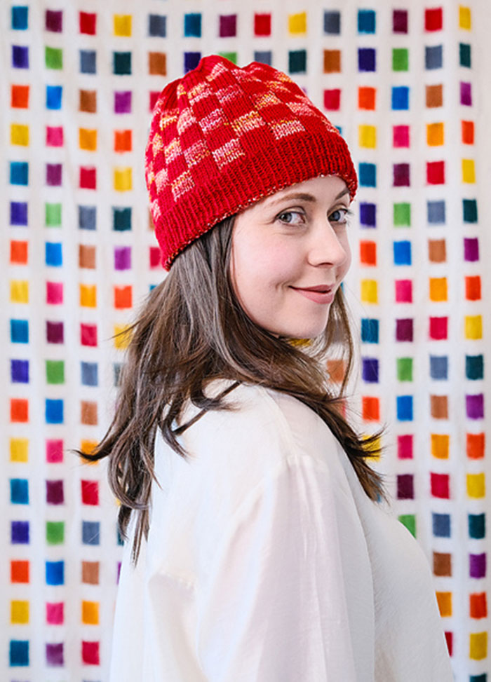 Side view of a Red Planet Hat with multicoloured checkered design, worn against a vibrant square-patterned backdrop.