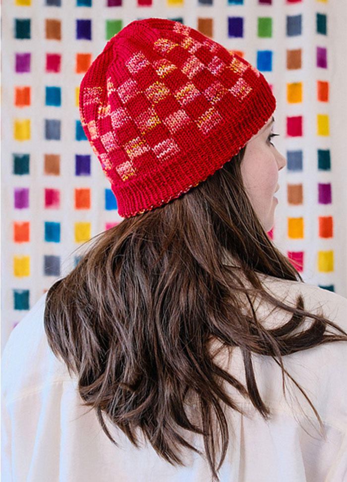 Angled view of a Red Planet Hat with geometric colourwork, shown in front of a colourful wall.