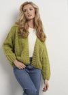 A woman with long blonde hair is wearing a chunky green knit cardigan over a white top and blue jeans. She stands against a textured white wall, looking contemplative.