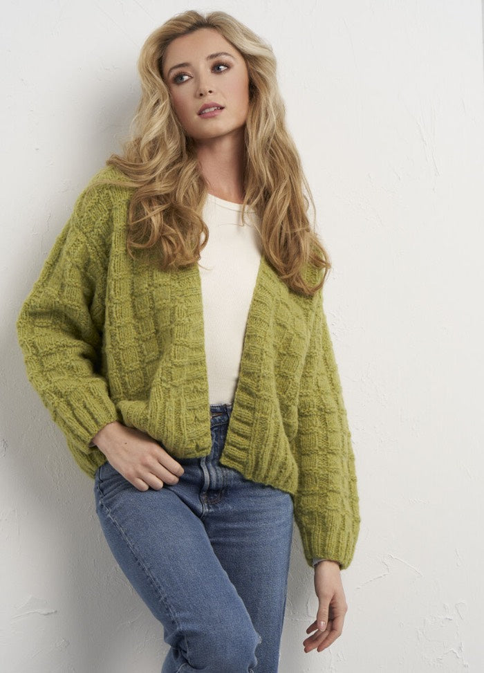 A woman with long blonde hair is wearing a chunky green knit cardigan over a white top and blue jeans. She stands against a textured white wall, looking contemplative.
