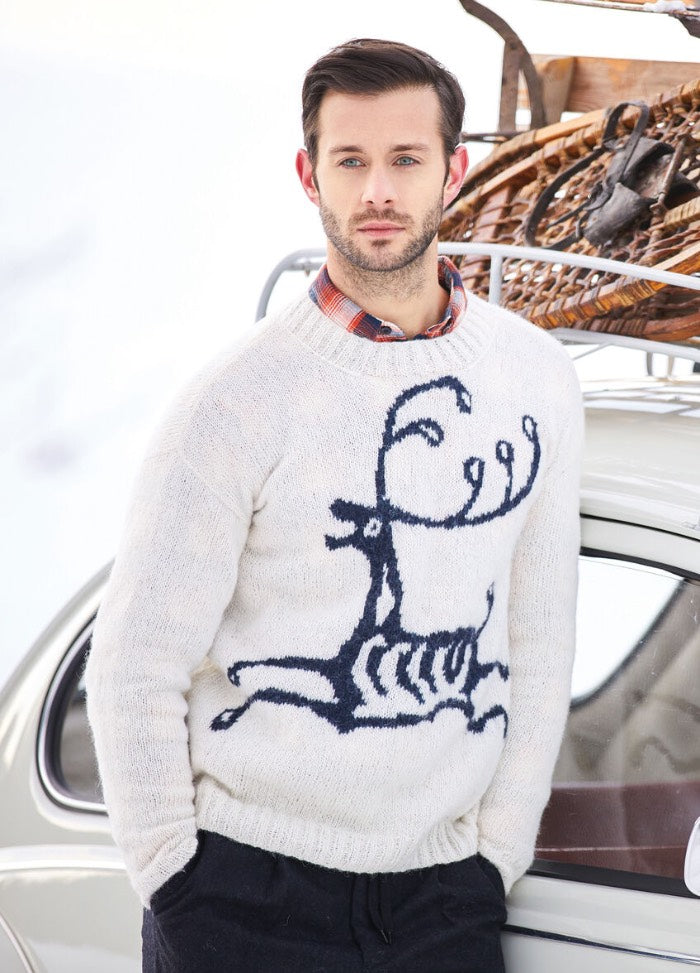 A man stands beside a vintage car in a snowy setting, wearing a white reindeer sweater and plaid shirt. Wooden sleds are on the car roof, adding a winter vibe.