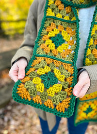 Retro Cardo Scarf held outdoors, crocheted in granny squares with green, yellow, and orange bordered in dark green.
