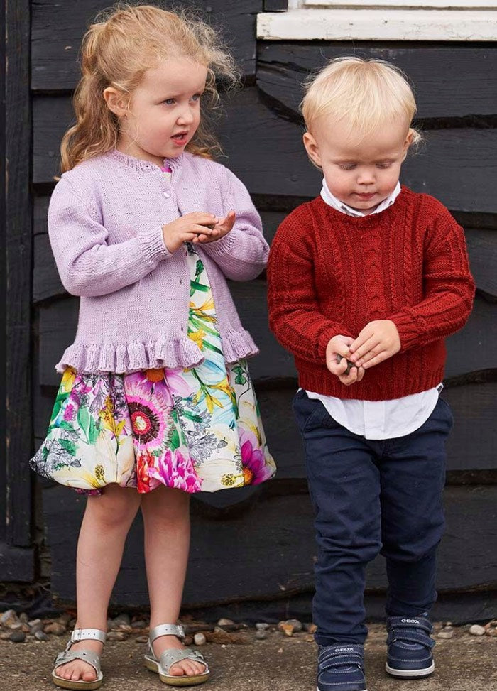 Two children stand outside against a dark wooden wall. The girl wears a purple cardigan and floral dress, the boy a red sweater and blue pants, both look curious.