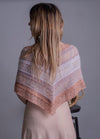 A woman with long hair is seen from behind, wearing a textured knit shawl in soft pink and white hues over a light dress against a neutral background.