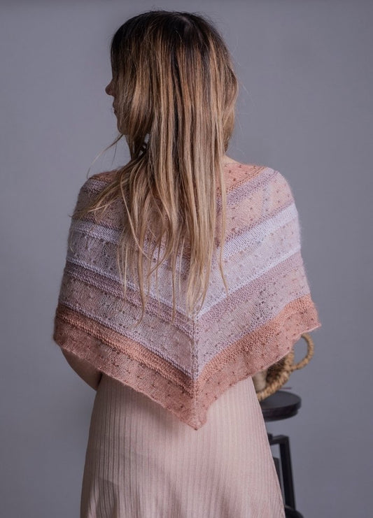 A woman with long hair is seen from behind, wearing a textured knit shawl in soft pink and white hues over a light dress against a neutral background.