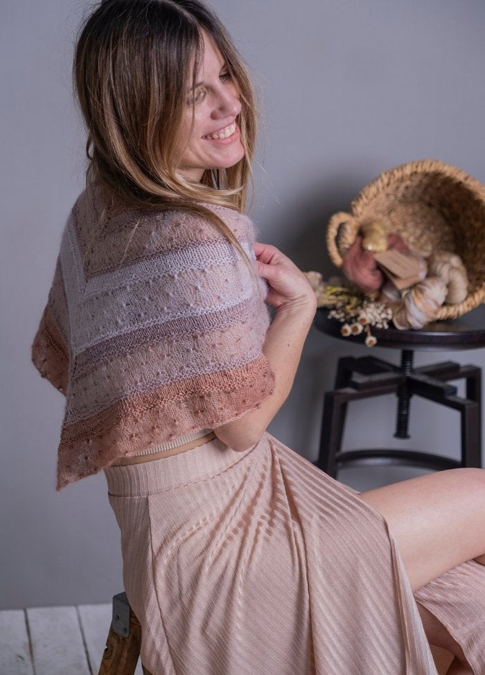 Smiling woman with long hair sits on a stool, wearing a delicate knit shawl and pleated skirt. A wicker basket with yarn is on a stool in the background.