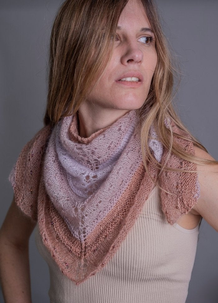 Woman in a beige tank top wears a soft pink and peach knitted lace shawl. She gazes to the side with a thoughtful expression. Neutral background.