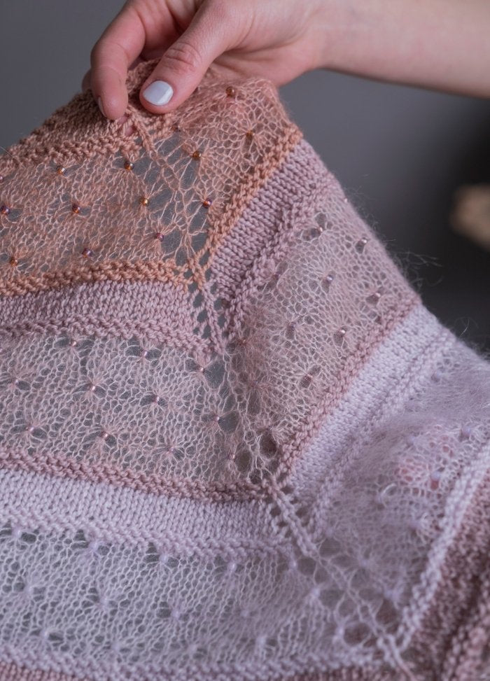 A hand holds a delicate knitted shawl in soft pink and peach tones, featuring intricate lace patterns and small beads, conveying a gentle, elegant feeling.