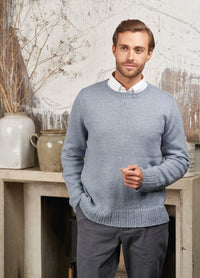 A man wearing a textured blue knit sweater and grey pants, standing confidently with a neutral background.