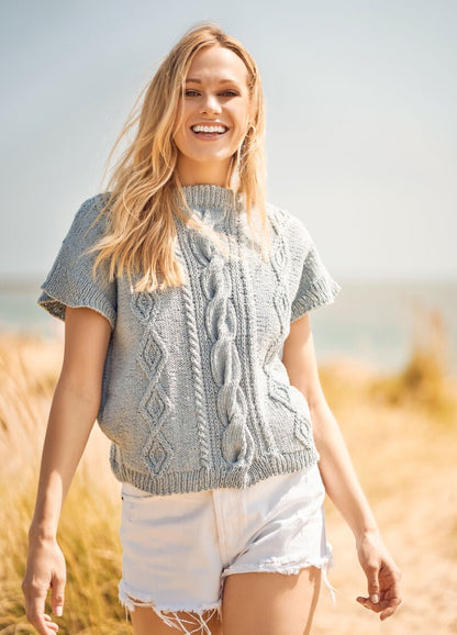 Smiling woman in a blue knit sweater and white shorts walking through a sunny beach landscape. Bright, cheerful mood with golden grass in the background.