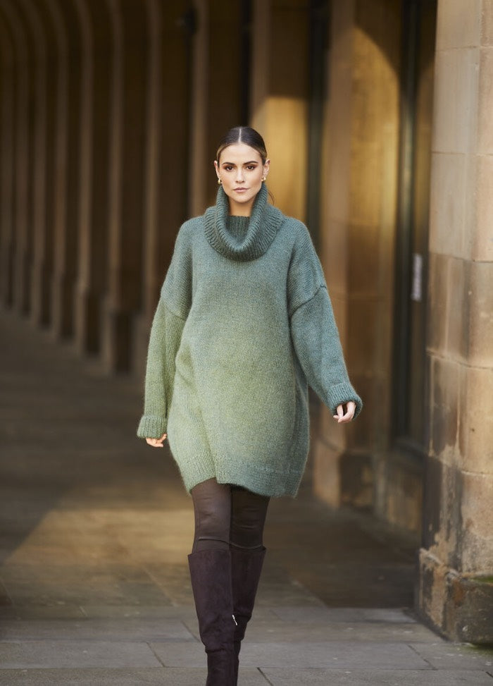 Woman in a teal oversized sweater and dark knee-high boots walks confidently down a stone corridor with soft, warm lighting. Urban, fashion-forward.