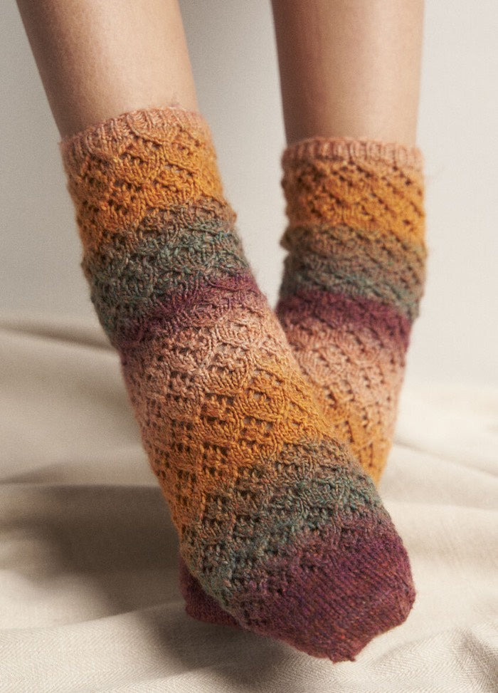 Cozy knitted socks feature an orange, green, and purple ombre pattern. Worn on crossed feet, the socks create a warm, textured feel against a soft background.