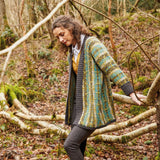 A woman in a knitted cardigan called Airadale by Rowan stands surrounded by lush greenery in a peaceful forest environment.