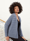 A person with curly hair wears a textured blue cardigan over a black outfit, posing confidently on a staircase with ornate railing. Neutral expression.